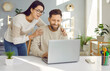© Studio Romantic - Happy young family couple using a laptop. Loving, supportive woman standing by her husband who is working on his notebook at his desk, holding him by the shoulders, looking at the computer and smiling