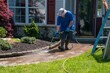 A caucasian man with a latex holding water sprayer wand power washing the brick walkway to a house