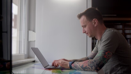 young guy with tattoos start work from home, sits down and open laptop. web designer or programmer at home office with refrigerator in background. man work in front of large window at home.