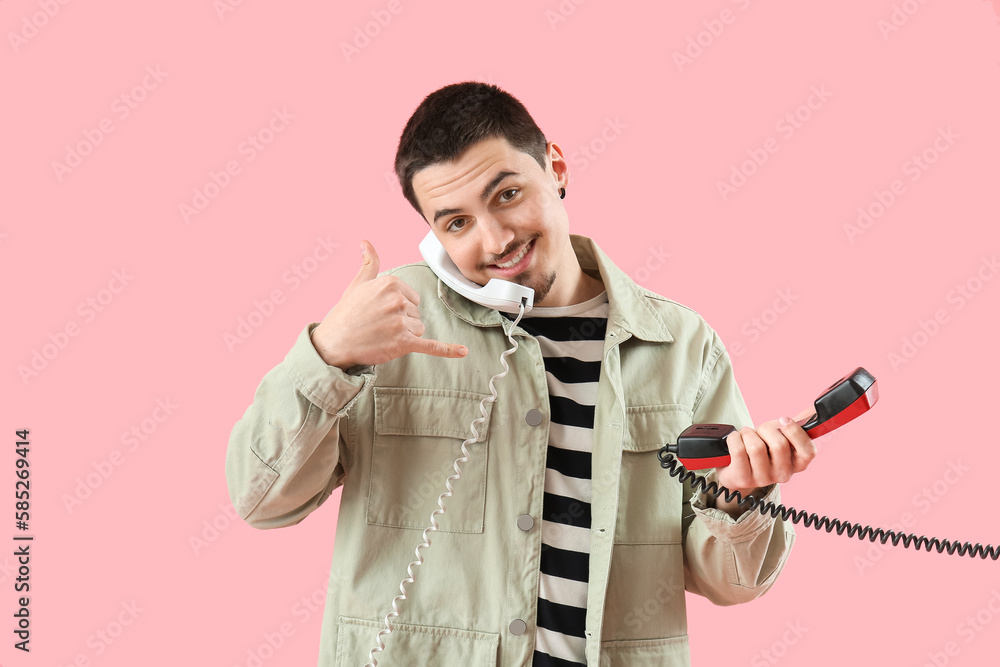 Cool young man with two phone receivers showing "call me" gesture on pink background