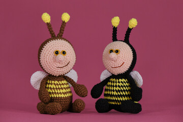 Amigurumi bumblebee dolls sit together on pink background. Soft DIY toy made of cotton. Two bees brown and black with yellow stripes and funny wings and horns crocheted, handmade art.