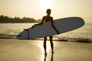 African american woman walking with surfboard on ocean beach. Black female surfer posing with surf board. Pretty multiethnic girl goes on surfing session, enjoys sun at tropical location at sunrise.