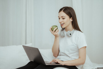 Wall Mural - Happy woman holding apple and listening to music sitting on sofa in living room at home