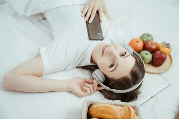 Wall Mural - Happy woman holding apple and listening to music sitting on sofa in living room at home