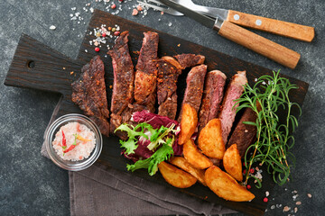 Steaks. Sliced grilled meat steak New York, Ribeye or Chuck roll with with garnished with salad and french fries on black marble board on old wooden background. Top view. Mock up.