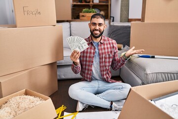 Canvas Print - Middle east man with beard sitting on the floor at new home holding money celebrating victory with happy smile and winner expression with raised hands