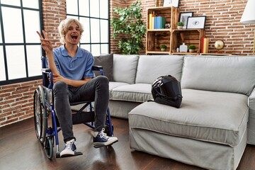 Poster - Young man sitting on wheelchair at for motorcycle accident smiling with happy face winking at the camera doing victory sign. number two.