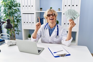 Canvas Print - Young doctor man holding money smiling with an idea or question pointing finger with happy face, number one