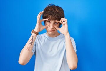 Sticker - Hispanic young man standing over blue background trying to open eyes with fingers, sleepy and tired for morning fatigue