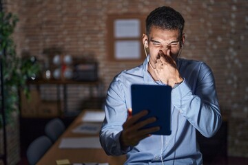 Poster - Handsome hispanic man working at the office at night smelling something stinky and disgusting, intolerable smell, holding breath with fingers on nose. bad smell