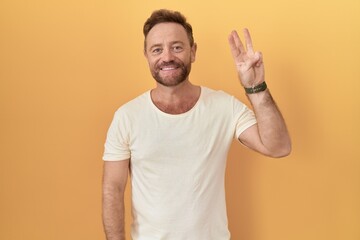 Sticker - Middle age man with beard standing over yellow background showing and pointing up with fingers number three while smiling confident and happy.