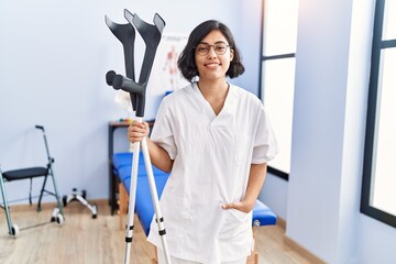 Poster - Young latin woman wearing physiotherapist uniform holding crutches at physiotherapy clinic