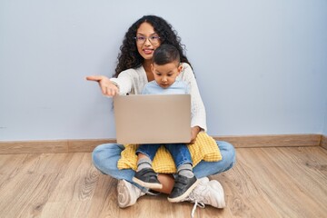 Poster - Young hispanic mother and kid using computer laptop sitting on the floor smiling cheerful offering palm hand giving assistance and acceptance.