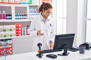 Poster - Young woman pharmacist holding pills bottle using computer at pharmacy