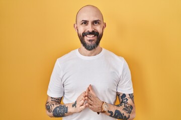 Poster - Young hispanic man with tattoos standing over yellow background hands together and fingers crossed smiling relaxed and cheerful. success and optimistic