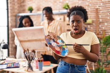 Sticker - Young black painter woman at art studio holding palette afraid and shocked with surprise and amazed expression, fear and excited face.