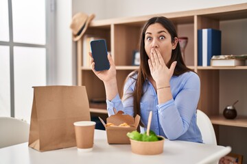 Canvas Print - Young brunette woman eating take away food at home showing smartphone screen covering mouth with hand, shocked and afraid for mistake. surprised expression