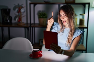 Sticker - Young hispanic woman using touchpad sitting on the table at night pointing finger up with successful idea. exited and happy. number one.