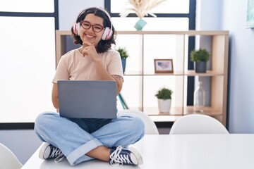 Sticker - Young hispanic woman using laptop sitting on the table wearing headphones looking confident at the camera smiling with crossed arms and hand raised on chin. thinking positive.