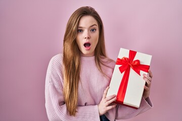 Canvas Print - Young caucasian woman holding gift afraid and shocked with surprise and amazed expression, fear and excited face.