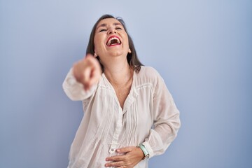 Wall Mural - Middle age hispanic woman standing over blue background laughing at you, pointing finger to the camera with hand over body, shame expression