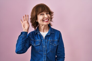 Wall Mural - Middle age woman standing over pink background waiving saying hello happy and smiling, friendly welcome gesture