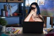 Young modern girl with blue hair sitting at art studio with laptop at night smelling something stinky and disgusting, intolerable smell, holding breath with fingers on nose. bad smell