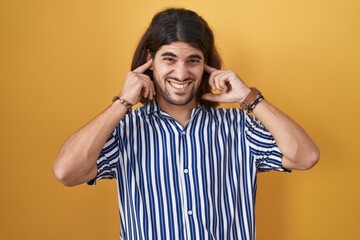 Wall Mural - Hispanic man with long hair standing over yellow background covering ears with fingers with annoyed expression for the noise of loud music. deaf concept.