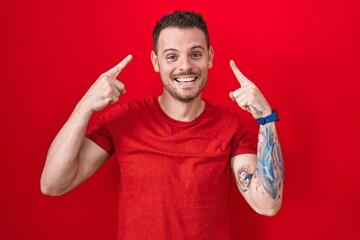 Canvas Print - Young hispanic man standing over red background smiling pointing to head with both hands finger, great idea or thought, good memory