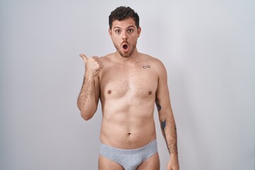 Wall Mural - Young hispanic man standing shirtless wearing underware surprised pointing with hand finger to the side, open mouth amazed expression.