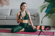 © ViDi Studio - Full body young strong athletic fitness trainer instructor woman wear green tracksuit earphones sit on floor use mobile cell phone drink water training do exercises at home gym Workout sport concept.