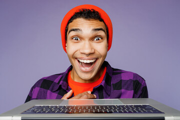 Close up young surprised student IT man of African American ethnicity wear casual shirt orange hat hold use work on laptop pc computer isolated on plain pastel purple color background studio portrait.