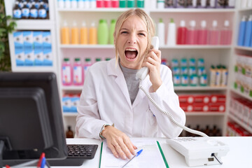 Poster - Young caucasian woman working at pharmacy drugstore speaking on the telephone angry and mad screaming frustrated and furious, shouting with anger. rage and aggressive concept.