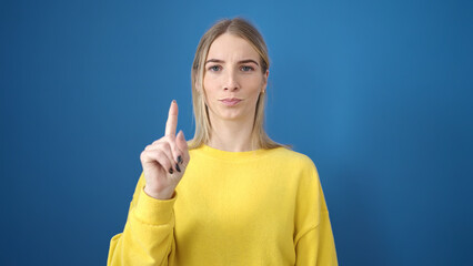 Sticker - Young blonde woman standing with serious expression pointing finger over isolated blue background