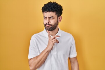 Wall Mural - Arab man standing over yellow background thinking concentrated about doubt with finger on chin and looking up wondering