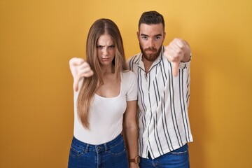 Wall Mural - Young couple standing over yellow background looking unhappy and angry showing rejection and negative with thumbs down gesture. bad expression.