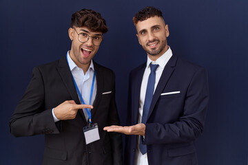 Poster - Two hispanic business men wearing business clothes amazed and smiling to the camera while presenting with hand and pointing with finger.