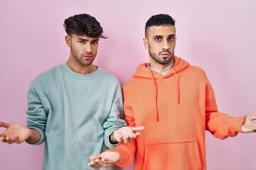 Wall Mural - Young hispanic gay couple standing over pink background clueless and confused with open arms, no idea concept.