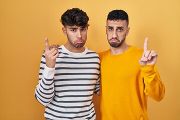 Poster - Young hispanic gay couple standing over yellow background pointing up looking sad and upset, indicating direction with fingers, unhappy and depressed.