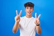 © Krakenimages.com - Hispanic teenager standing over blue background smiling with tongue out showing fingers of both hands doing victory sign. number two.