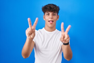 Canvas Print - Hispanic teenager standing over blue background smiling with tongue out showing fingers of both hands doing victory sign. number two.