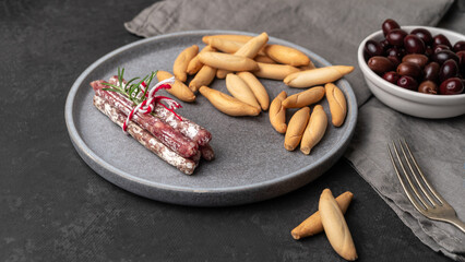 Close-up of the Plate with Spanish sticks of mini fuet and picos - bread sticks and bowl with dark olives on the grey background.