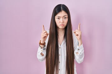 Poster - Chinese young woman standing over pink background pointing up looking sad and upset, indicating direction with fingers, unhappy and depressed.