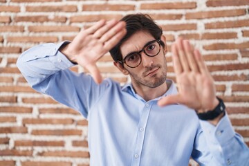 Poster - Young hispanic man standing over brick wall background doing frame using hands palms and fingers, camera perspective