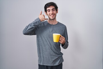 Poster - Young hispanic man wearing pajama drinking a cup of coffee smiling doing phone gesture with hand and fingers like talking on the telephone. communicating concepts.