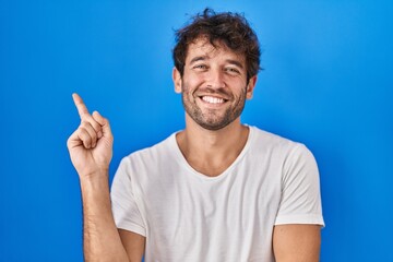 Canvas Print - Hispanic young man standing over blue background with a big smile on face, pointing with hand finger to the side looking at the camera.