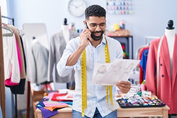 Poster - Young arab man tailor talking on smartphone looking clothing design at tailor shop