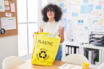 Sticker - Young middle east woman holding recycling bag with plastic bottles at the office smiling looking to the side and staring away thinking.