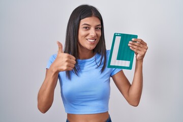 Brunette young woman holding l sign for new driver smiling happy and positive, thumb up doing excellent and approval sign