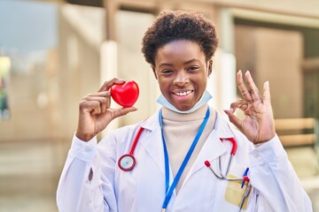 Poster - African american woman wearing doctor uniform holding heart doing ok sign with fingers, smiling friendly gesturing excellent symbol